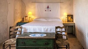Una habitación con cama, dos mesitas de noche, un aparador verde, dos sillas de mimbre y un tapiz de pared.
