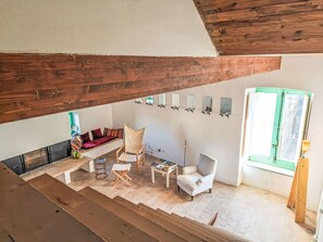 Ein Loft-Zimmer mit hohen Decken, einer integrierten Sitzecke, einem kleinen Tisch mit Büchern und einem Fenster mit grünem Rahmen.