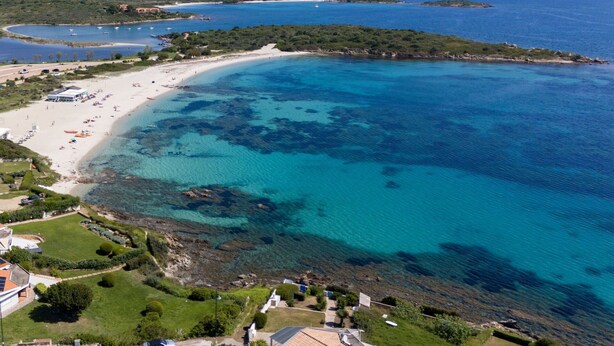 An aerial view of a coastal area with a sandy beach, clear blue waters, and a small peninsula with buildings.