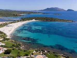 Vista aérea de uma área costeira com praia de areia, águas azul-claras e uma pequena península com edifícios.