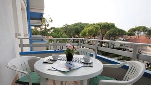 A small outdoor balcony with a round table, two chairs, a potted plant, and a view of trees and buildings.