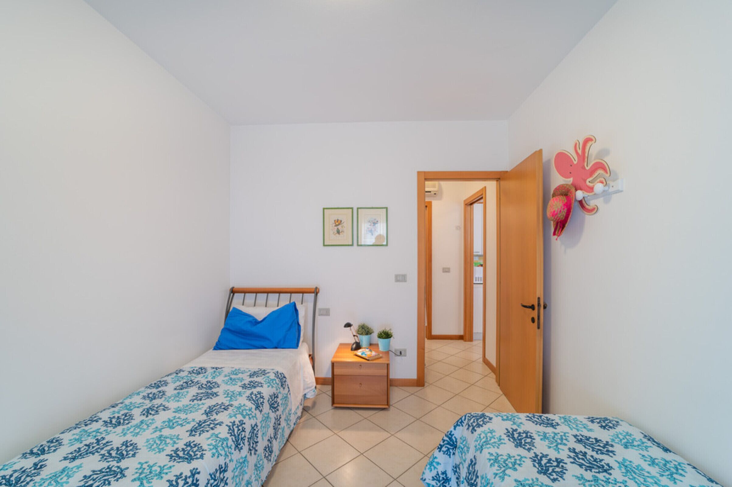 Une petite chambre propre, avec deux lits simples, une table de chevet, une petite table et une porte donnant sur une autre pièce.