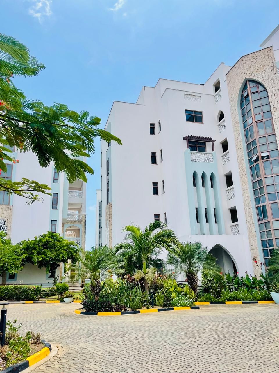 Un edificio de varios pisos con un diseño arquitectónico moderno, que presenta una combinación de ventanas planas y arqueadas, y un patio con palmeras y plantas en macetas.