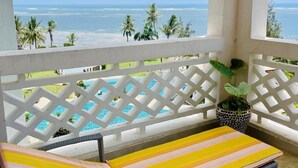 A balcony with a view of the ocean, a pool, and a wicker lounge chair with a striped cushion. - Luxury Oceanline Apartments-Shaza. (Mombasa)