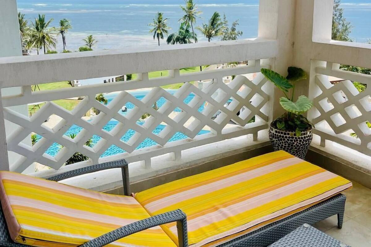 A balcony with a view of the ocean, a pool, and a wicker lounge chair with a striped cushion.