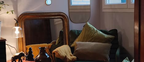 A cozy living room with a green sofa, a small table with decorative items, a mirror, and a window with a view of trees.