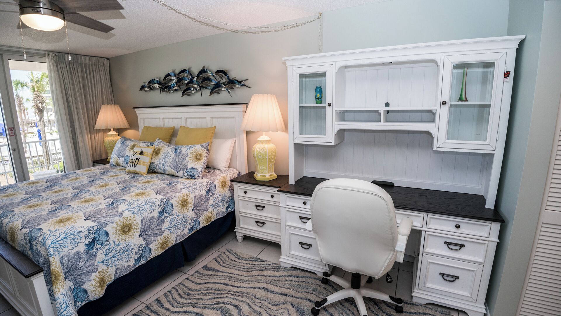 A bedroom with a bed, a desk, and a chair. There is a window with curtains, a ceiling fan, and a wall-mounted shelf with decorative items.