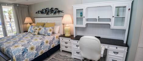 A bedroom with a bed, a desk, and a chair. There is a window with curtains, a ceiling fan, and a wall-mounted shelf with decorative items.