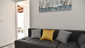 Un salon moderne avec un canapé d’angle gris, deux coussins, un tableau représentant des voiliers et une porte donnant sur un espace extérieur.