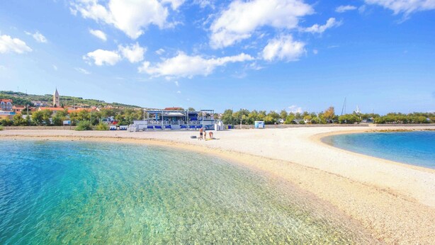Une plage avec une eau cristalline, un rivage sablonneux et un bâtiment surmonté d’un toit bleu.