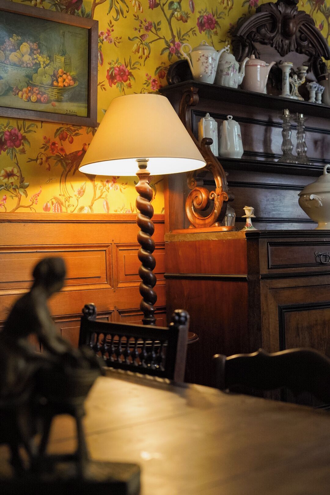 A wooden table with a lamp, a chair, and a decorative shelf with teapots and a framed painting.
