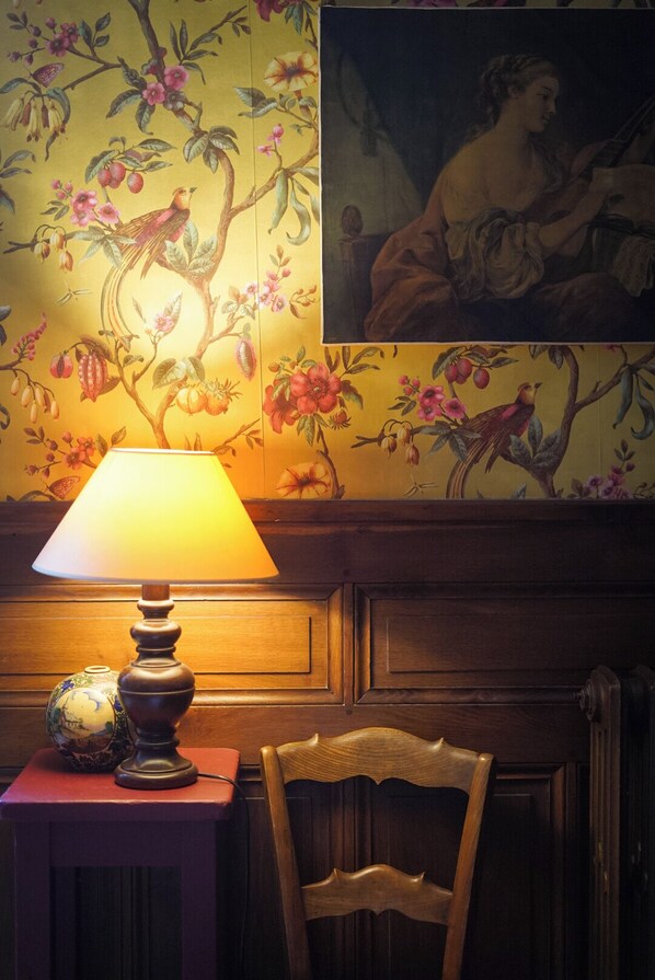 A wooden chair and a table with a lamp and a globe, against a wall with floral wallpaper and a framed painting. - La Maison Saint Aignan (Saint-Aignan)