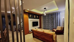 A modern living room with a large wooden sofa, a flat-screen TV mounted on the wall, and a ceiling light fixture. - Staycation At Sandari Batulao (Overlooking Spot) (Tagaytay)
