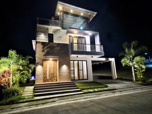 A modern two-story house with a wooden front door, a balcony on the upper floor, and a well-lit entrance. - Staycation At Sandari Batulao (Overlooking Spot) (Tagaytay)