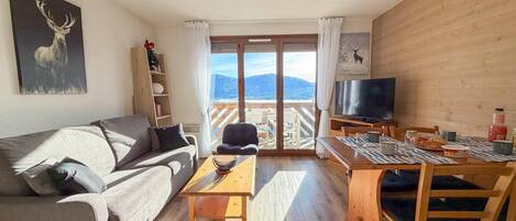 Un salón acogedor con sofá, mesa de centro, televisor y balcón con vistas a la montaña.