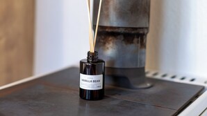 Eine schwarze Flasche mit dem Aufdruck "MOHEDA VANILLA BEAN", in die zwei Stangen gesteckt sind, auf einer Metalloberfläche in der Nähe eines großen, zylindrischen Objekts platziert.