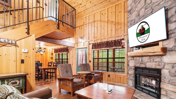 L’intérieur chaleureux d’un chalet, avec un décor en bois, une cheminée en pierre, un coin repas et une grande fenêtre donnant sur le paysage extérieur.