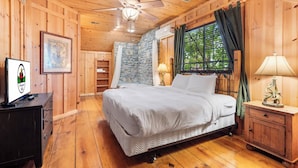Une chambre dans une cabane en bois, avec un lit, un ventilateur de plafond, un mur en pierre et une fenêtre avec des rideaux.