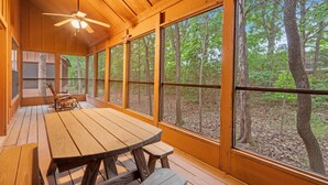 A wooden deck with a picnic table and benches, large windows offering forest views, and a ceiling fan.