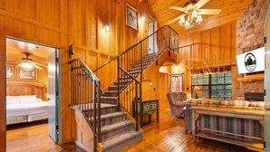 A wooden cabin interior with a staircase, a bed, a fireplace, and a dining area.