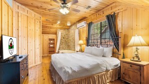 A wooden cabin bedroom with a bed, nightstands, a dresser, a ceiling fan, and a stone wall.
