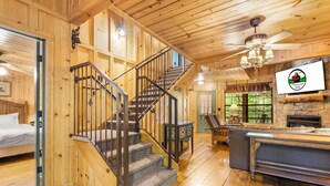 A wooden cabin interior with a staircase, a fireplace, a dining area, and a bed.