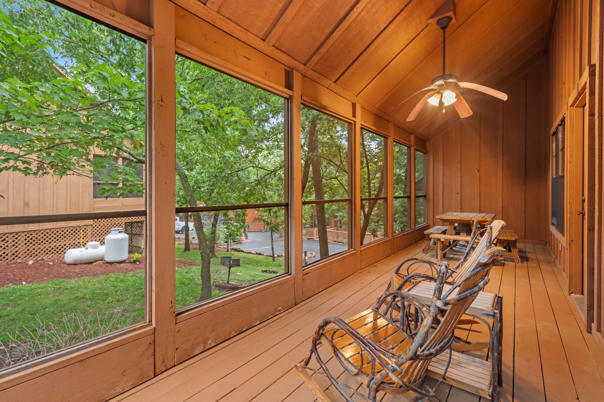 A covered outdoor seating area with wooden tables and chairs, large windows, and a ceiling fan.