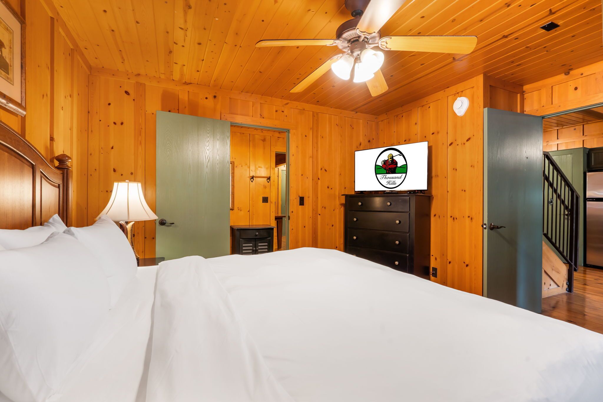 A wooden-paneled room with a bed, a ceiling fan, a dresser, and a television displaying a logo.