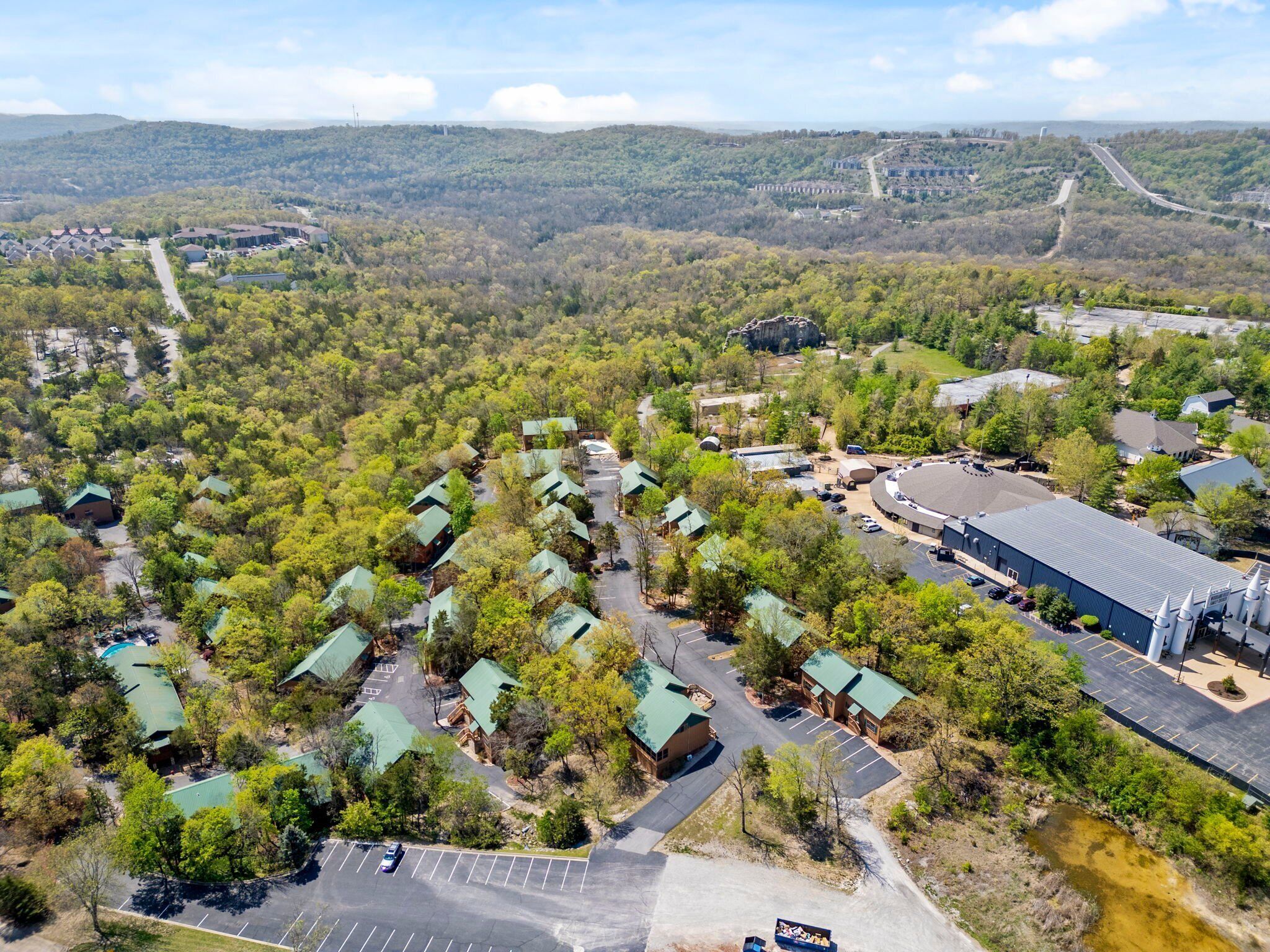An aerial view of a resort with multiple green-roofed cabins, a large building with a flat roof, and a parking area.