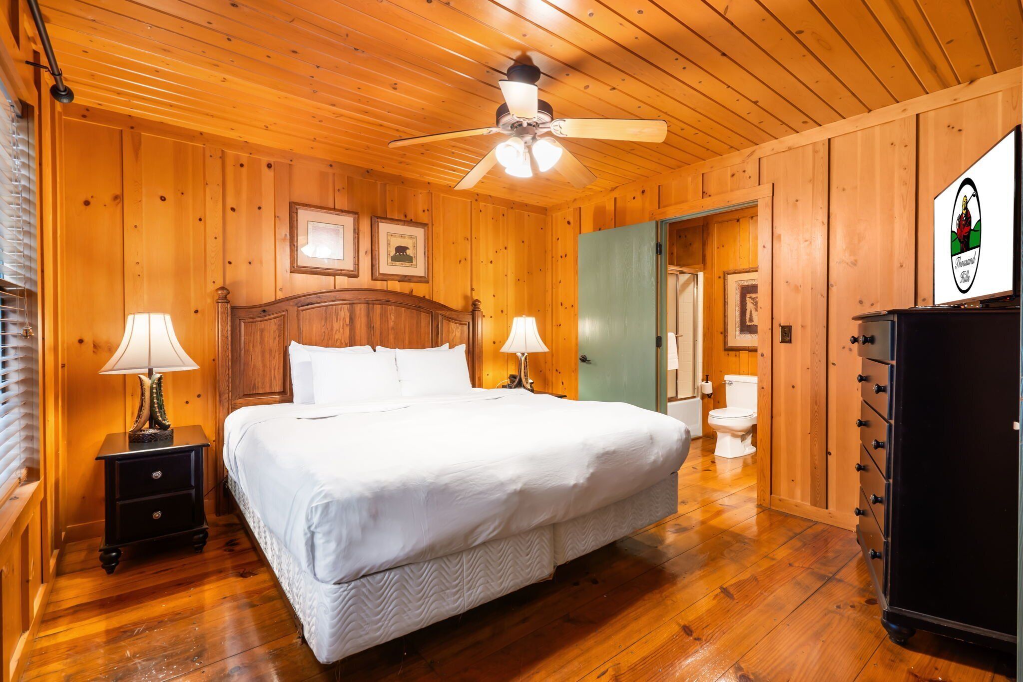 A wooden-paneled bedroom with a bed, two bedside lamps, a ceiling fan, and a dresser.