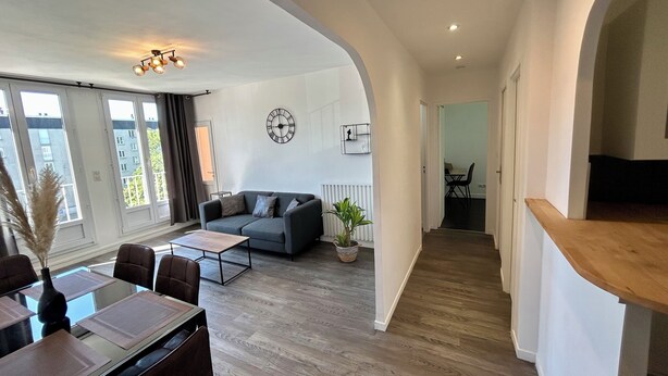 Un salon moderne avec un canapé gris, une table à manger avec des chaises et une grande fenêtre avec des rideaux.