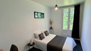 Une chambre à coucher comprenant un lit, une table de chevet, une lampe, une fenêtre avec des rideaux et un luminaire suspendu.