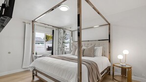 A bedroom with a four-poster canopy bed, a bedside table with a lamp, and a view of the outside through a window.