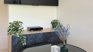 En moderne stue med et fladskærms-TV på væggen, et marmorbord med en computer, et rundt glasbord med en vase blomster og en potteplante.