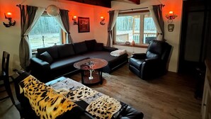 A cozy living room with a sectional sofa, a leather armchair, and a patterned ottoman. There are two windows with curtains, a wooden ceiling, and wall-mounted lamps.