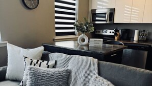 A modern living room with a grey sofa, a clock, and a kitchen in the background. - 12 Condo/King Bed/Close To  Attractions/Laundry (Niagara Falls)