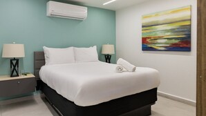 A modern hotel room with a bed, two bedside tables with lamps, an air conditioning unit, and a colorful abstract painting on the wall.