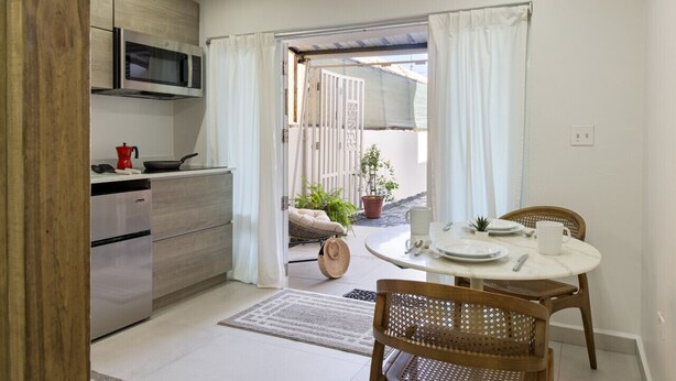 A compact kitchen with modern appliances, a dining area with a round table and chairs, and a view of a garden through an open door.