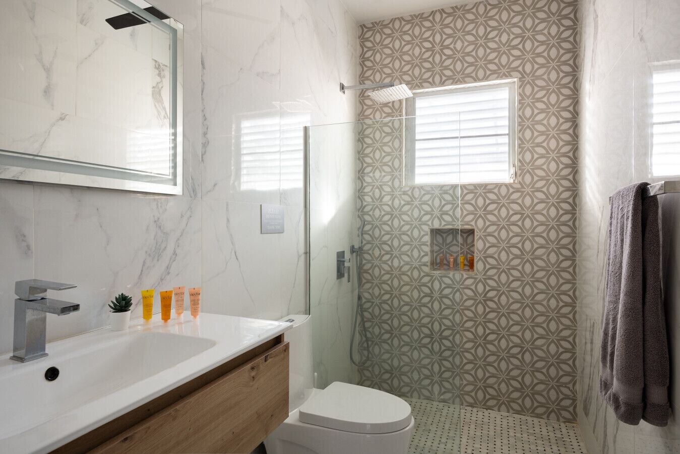 Une salle de bain avec un meuble-lavabo en marbre, une douche dont le mur est orné d’un motif géométrique et une petite fenêtre.