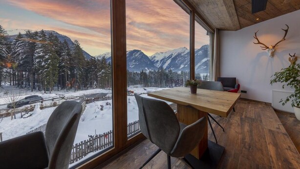 A room with a large window offering a view of snow-covered mountains and a forest. There is a wooden table and two chairs. - Chalet With Sauna and Natural Pool (Neukirchen am Grossvenediger)