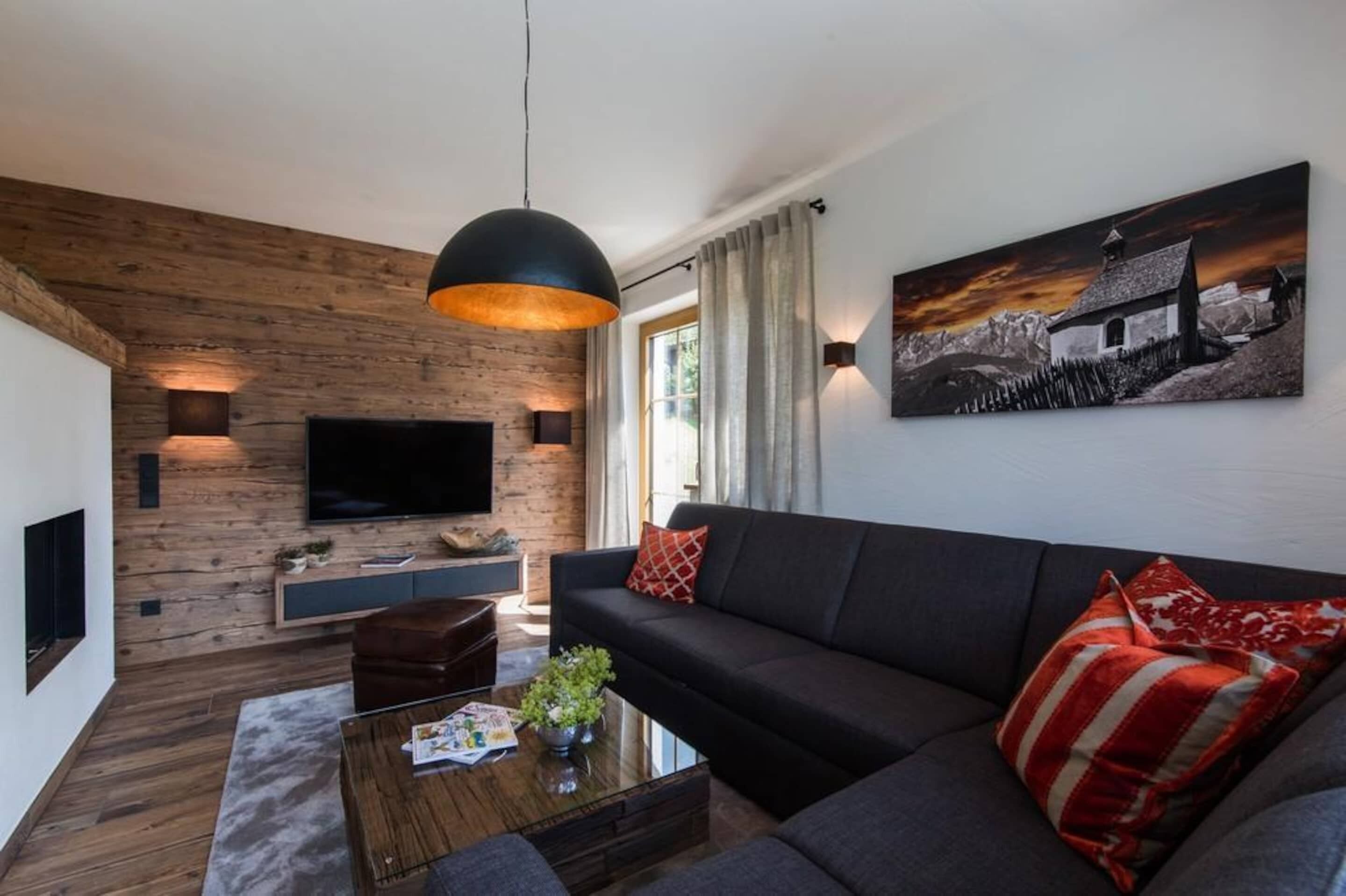 Ein modernes Wohnzimmer mit einer Wand als Holzdekor, einem großen Flachbildfernseher, einem dunklen Sofa und einem roten und weißen Kissen mit Muster.