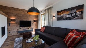 A modern living room with a wooden accent wall, a large flat-screen TV, a dark sofa, and a red and white patterned throw pillow. - Chalet With Sauna and Natural Pool (Neukirchen am Grossvenediger)