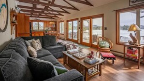 A cozy living room with a sectional sofa, wooden beams on the ceiling, a dining area with a table and chairs, and large windows offering a view of the outdoors.
