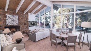 A cozy living room with a stone fireplace, a large window with a view of trees, and a dining area with a round table and chairs.