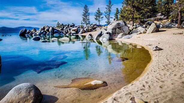 A clear lake with rocks and trees, a sandy beach, and a clear sky.