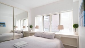 A modern hotel room with a large bed, two bedside lamps, a window with a view of buildings, and a mirror.