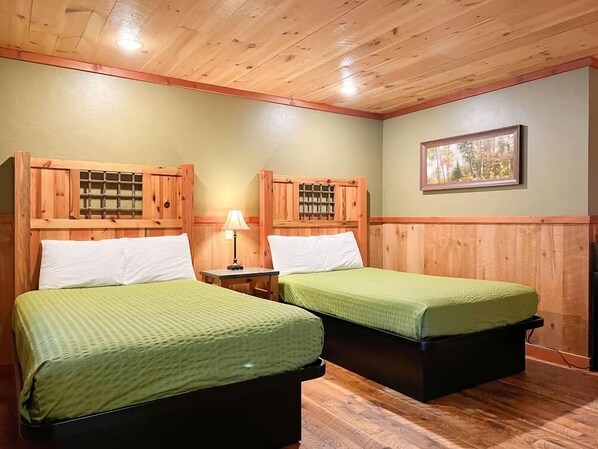 A room with two beds, wooden headboards, a small table with a lamp, and a framed picture on the wall.