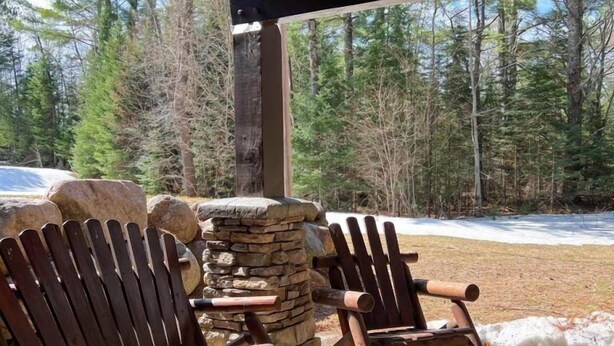 A covered outdoor seating area with Adirondack chairs and a stone table, overlooking a snowy landscape and dense forest. - Club 51 (Mercer)