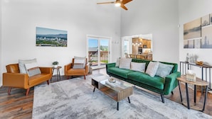 Ein geräumiges Wohnzimmer mit einem grünen Sofa, zwei Sesseln, einem Couchtisch und einem Deckenventilator. Ein Stadtbild hängt an der Wand, und durch eine Glastür ist ein Blick ins Freie möglich.