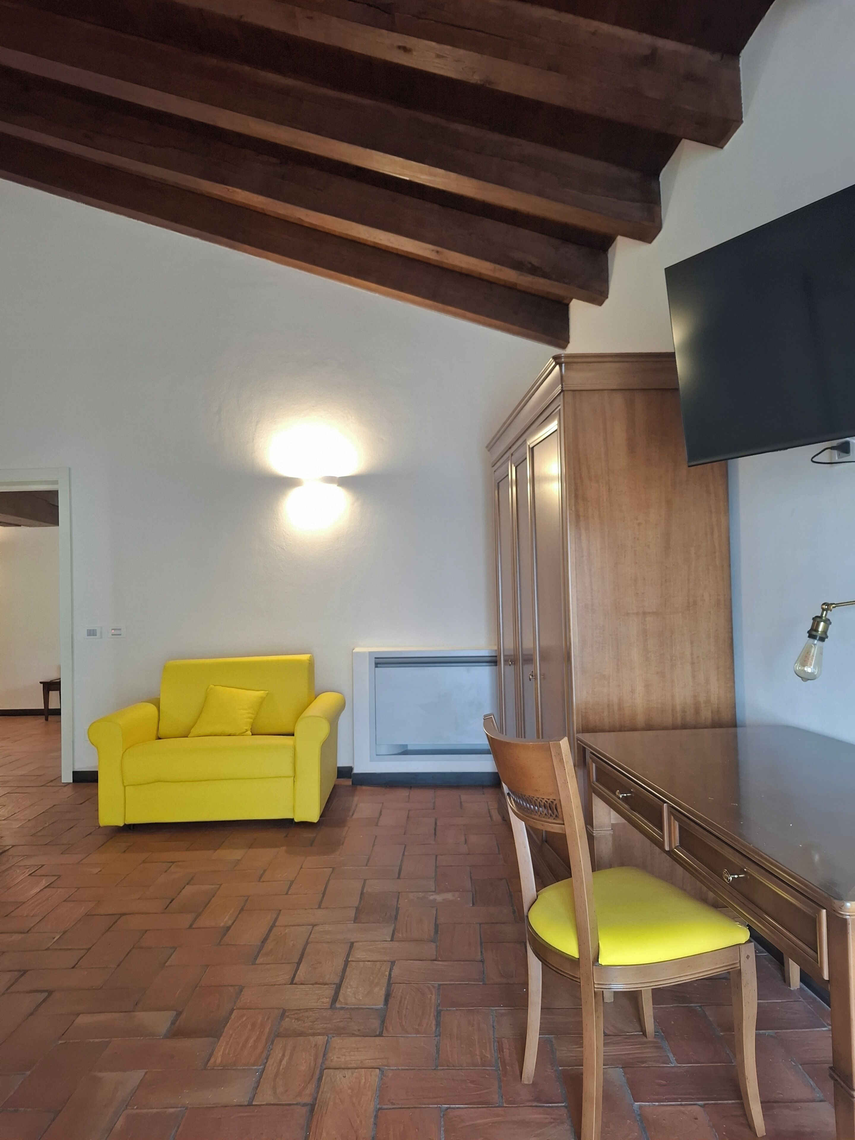A room with a wooden ceiling, a yellow sofa, a wooden chair, a TV mounted on the wall, and a radiator.